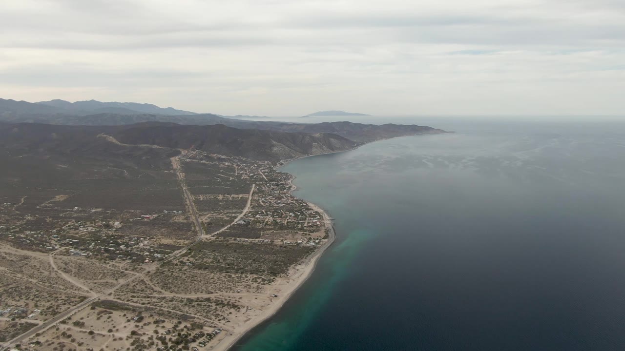 Los Barriles aerial view with the sandy coast at lower right, a compact settlement and road network to the left, dark hills behind town, broad ocean to the right, and no visible people.