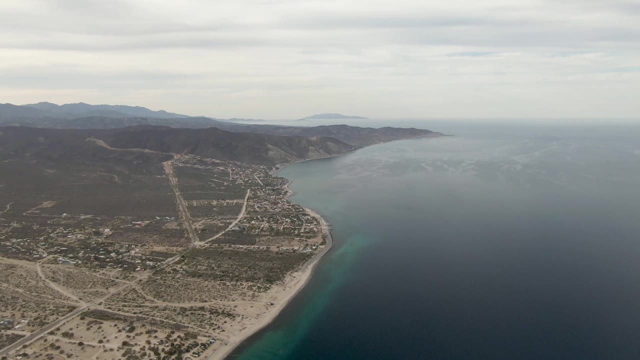 Mid-clip aerial frame over Los Barriles with more open water dominating the right side, sandy beach and scattered buildings along the shore, mountain ridges in the distance, and no visible people.