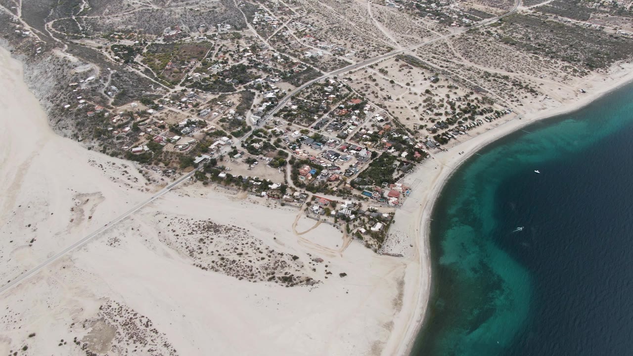Aerial frame over Los Barriles with the beach sweeping below the houses and the turquoise shallows concentrated near the right side, with no visible people.