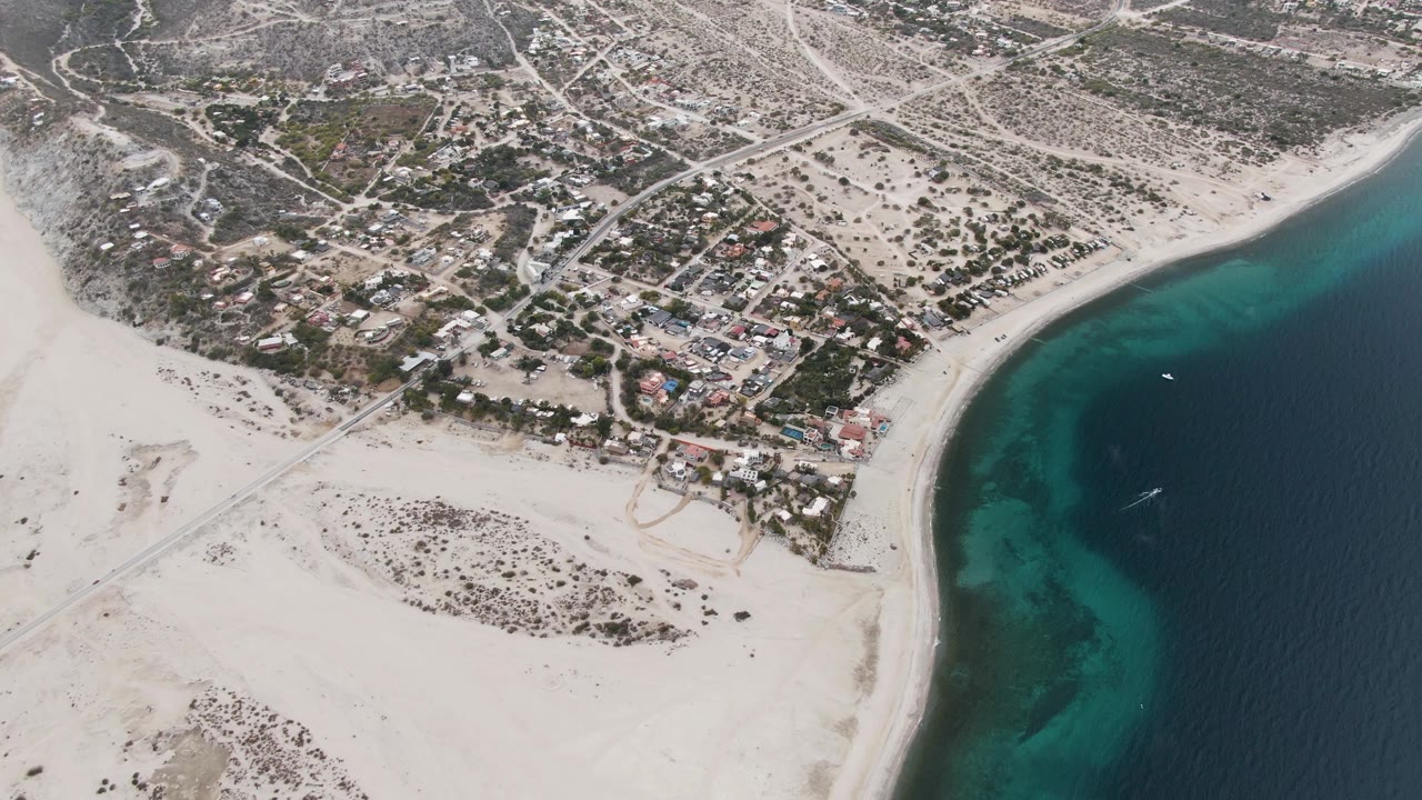High aerial frame over Los Barriles, Baja California Sur, showing a curving sandy beach, turquoise shallows, deeper blue water, inland roads, scattered homes, scrub terrain, and no visible people.
