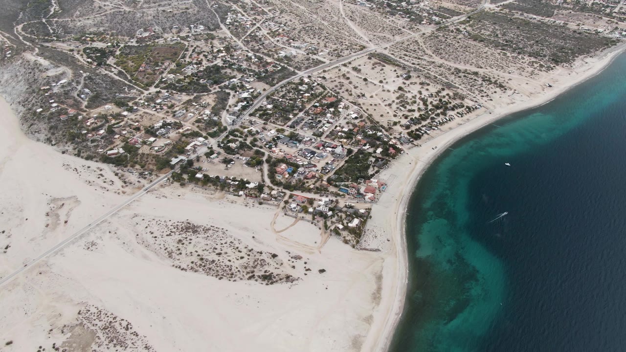 Aerial view in Los Barriles with the sandy shoreline running along the right side, compact residential blocks inland, dry terrain beyond the roads, a small boat offshore, and no visible people.