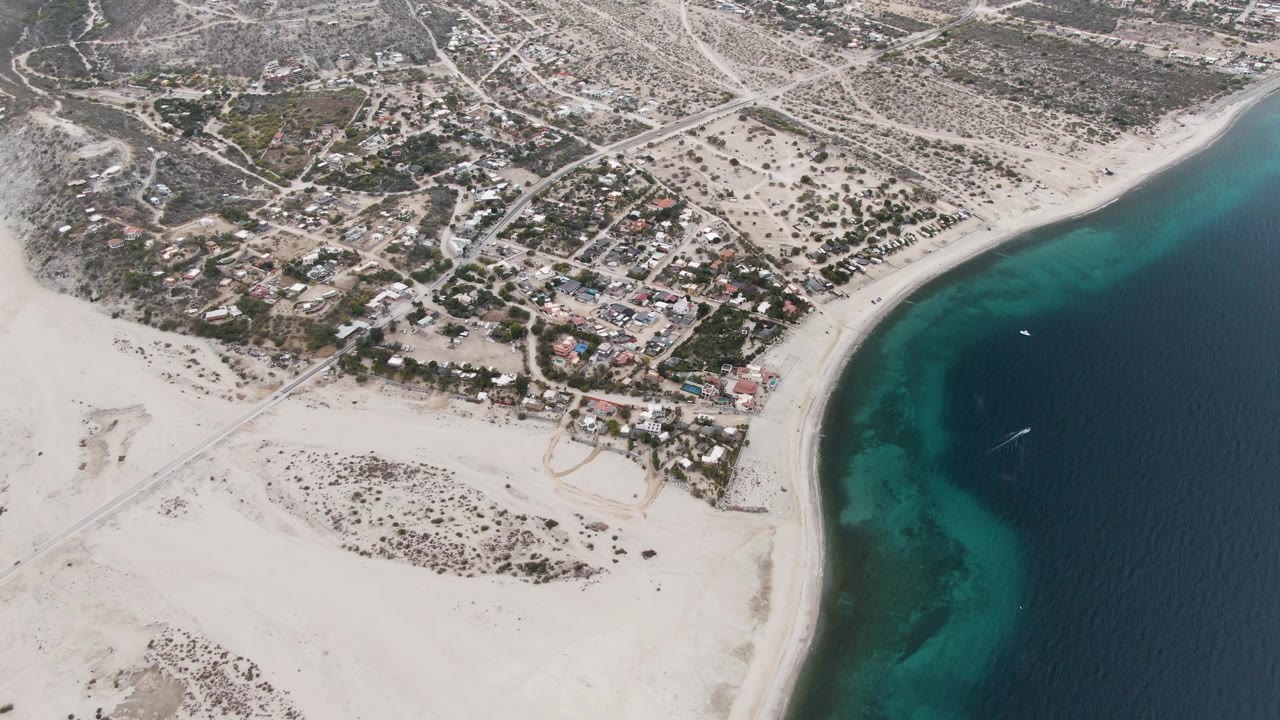 High oblique frame over Los Barriles showing the beach arc, turquoise nearshore water fading into dark blue ocean, inland streets, clustered homes, open sandy lots, and no visible people.