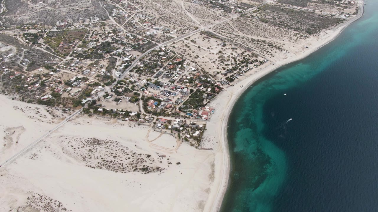 Aerial frame over Los Barriles, Baja California Sur, with pale sand in the foreground, scattered buildings and roads inland, turquoise shoreline at right, a small boat offshore, and no visible people.