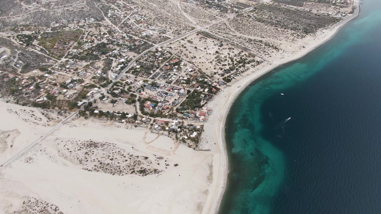 At 0:01, the Los Barriles coastline remains framed from above, with the curved beach separating dry inland lots from turquoise water and a small white boat slightly farther along shore, with no visible people.