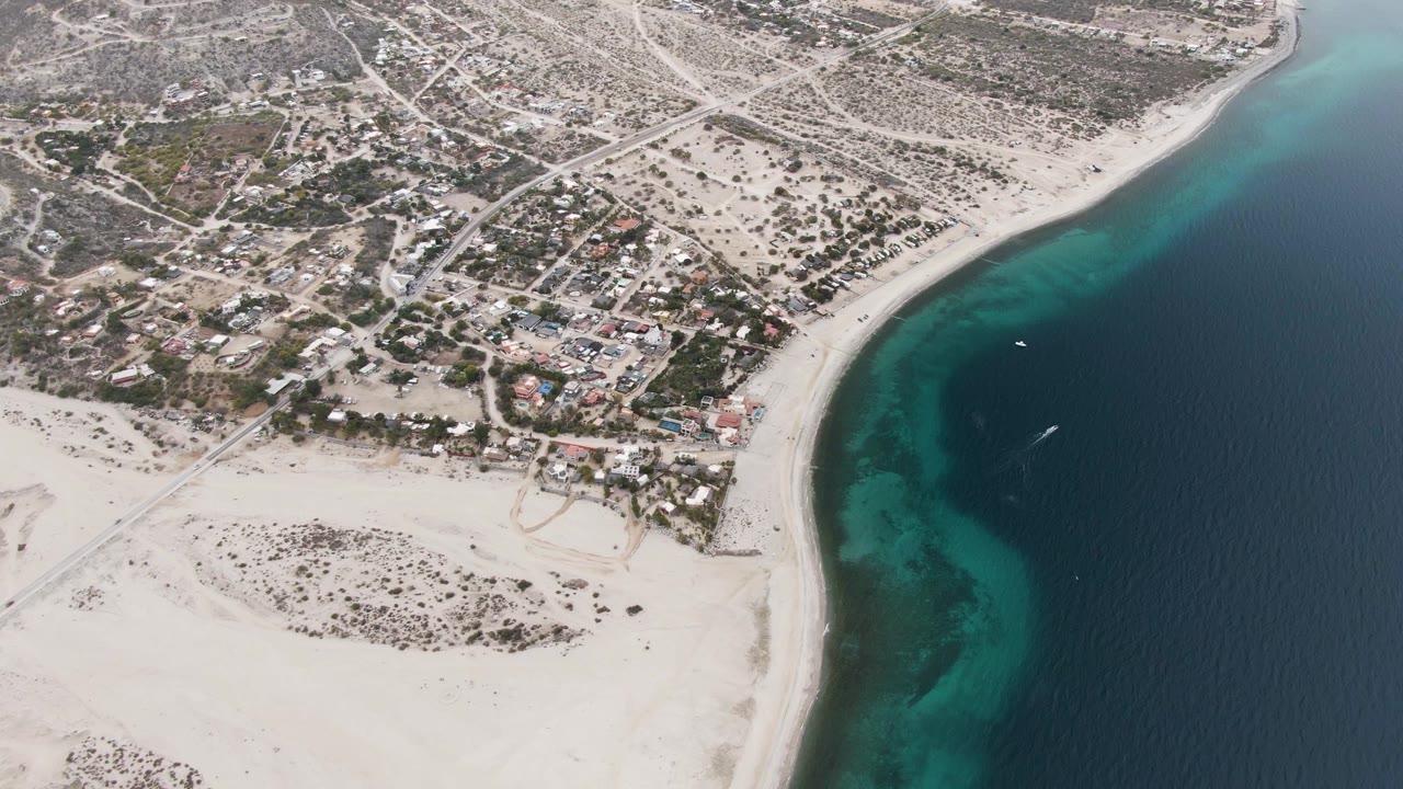 At 0:02, the aerial view of Los Barriles shows bright sand and shallow turquoise water beside darker ocean, with a grid of roads and scattered buildings inland and no visible people.