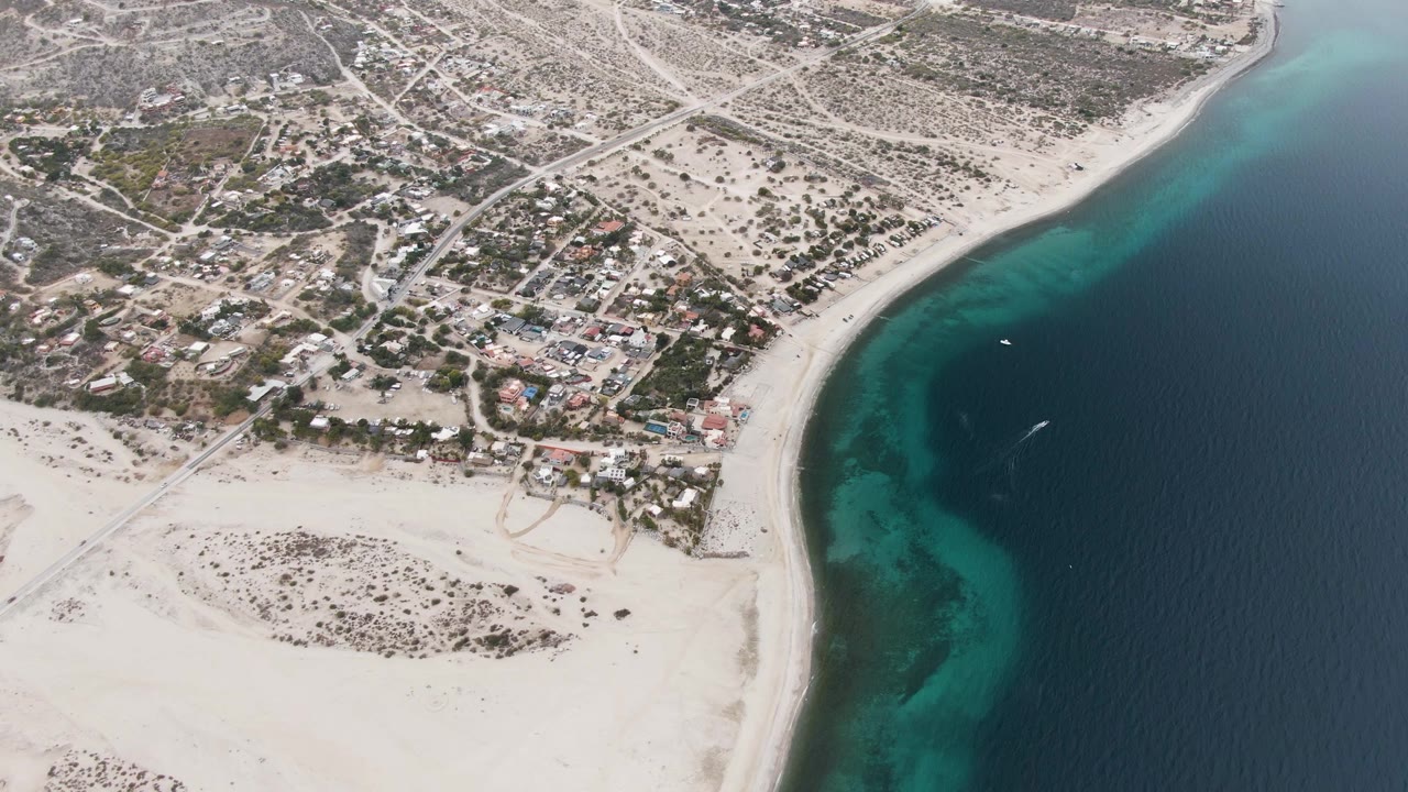 Aerial coastline view with sandy beach, settlement, and boat wake in blue water.