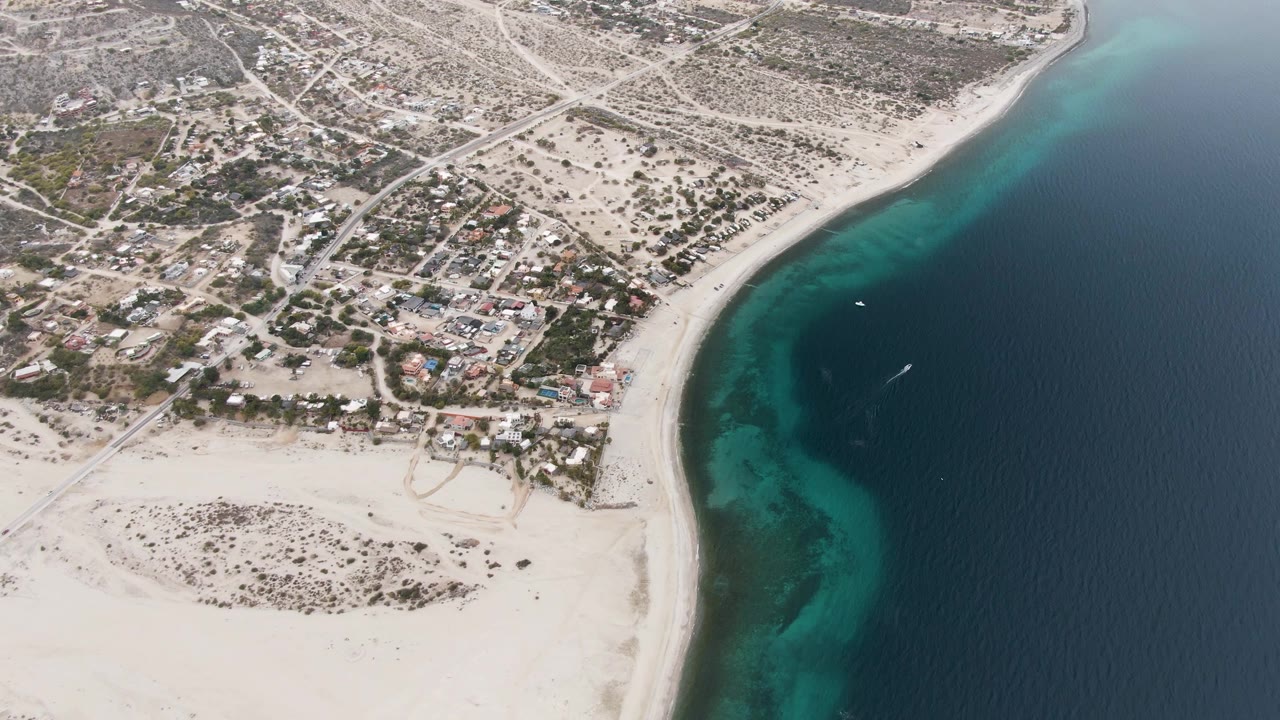 Opening aerial frame over Los Barriles, Baja California Sur, with dry residential land at left, a curving sandy beach, turquoise shallows, deep blue water, and no visible people.