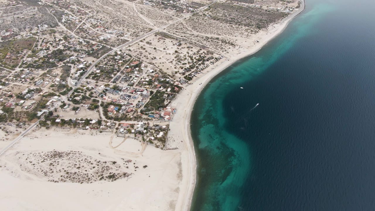 Third aerial frame from Los Barriles with the residential grid and pale arid ground at left, the beach running diagonally through center, blue-green shallows, and no visible people.