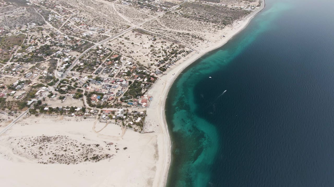 Aerial shoreline with dry land, homes, beach, and dark blue sea.