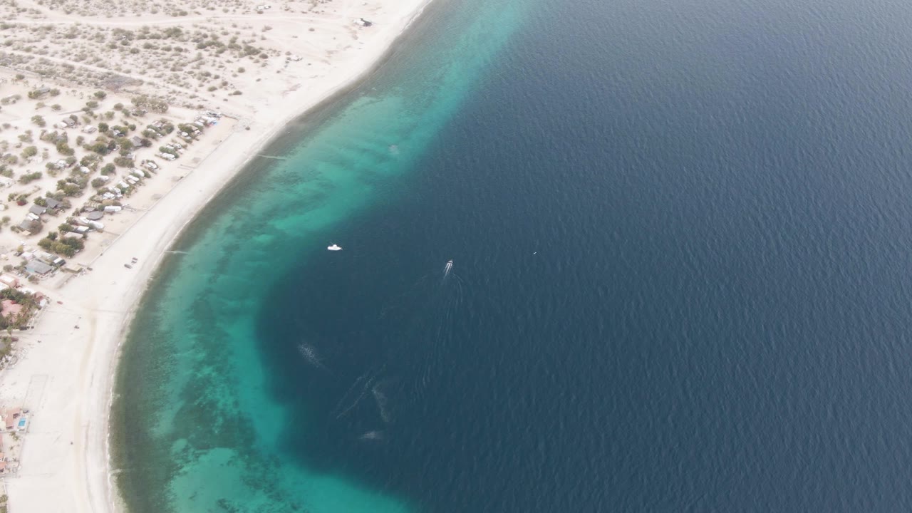 Aerial frame over Los Barriles showing a pale sandy beach curving beside turquoise shallows and darker open water, with no visible people.