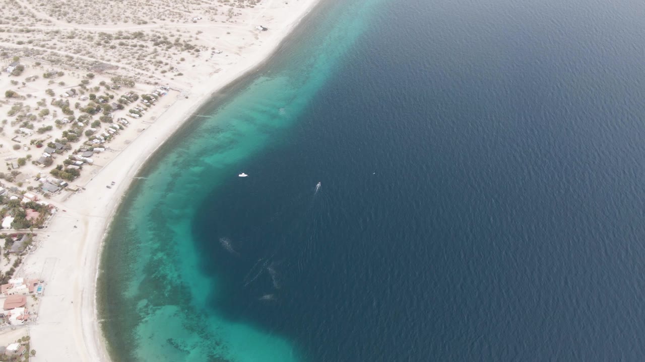 Drone view over Los Barriles with shoreline buildings on the sand, two small boats in the water, and turquoise shallows fading into deep blue, with no visible people.