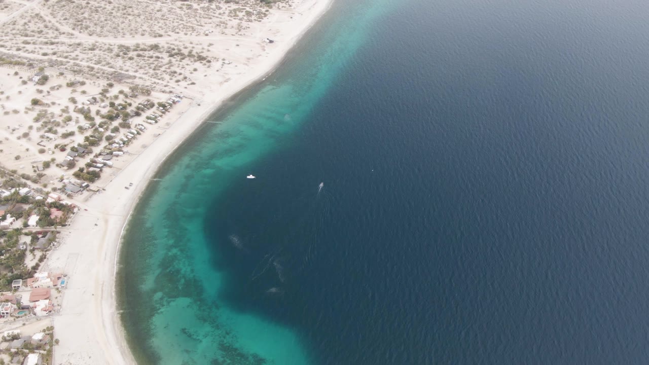 High aerial frame of Los Barriles coastline with a pale beach on the left, a bright turquoise nearshore band, and rippled dark blue water on the right, with no visible people.
