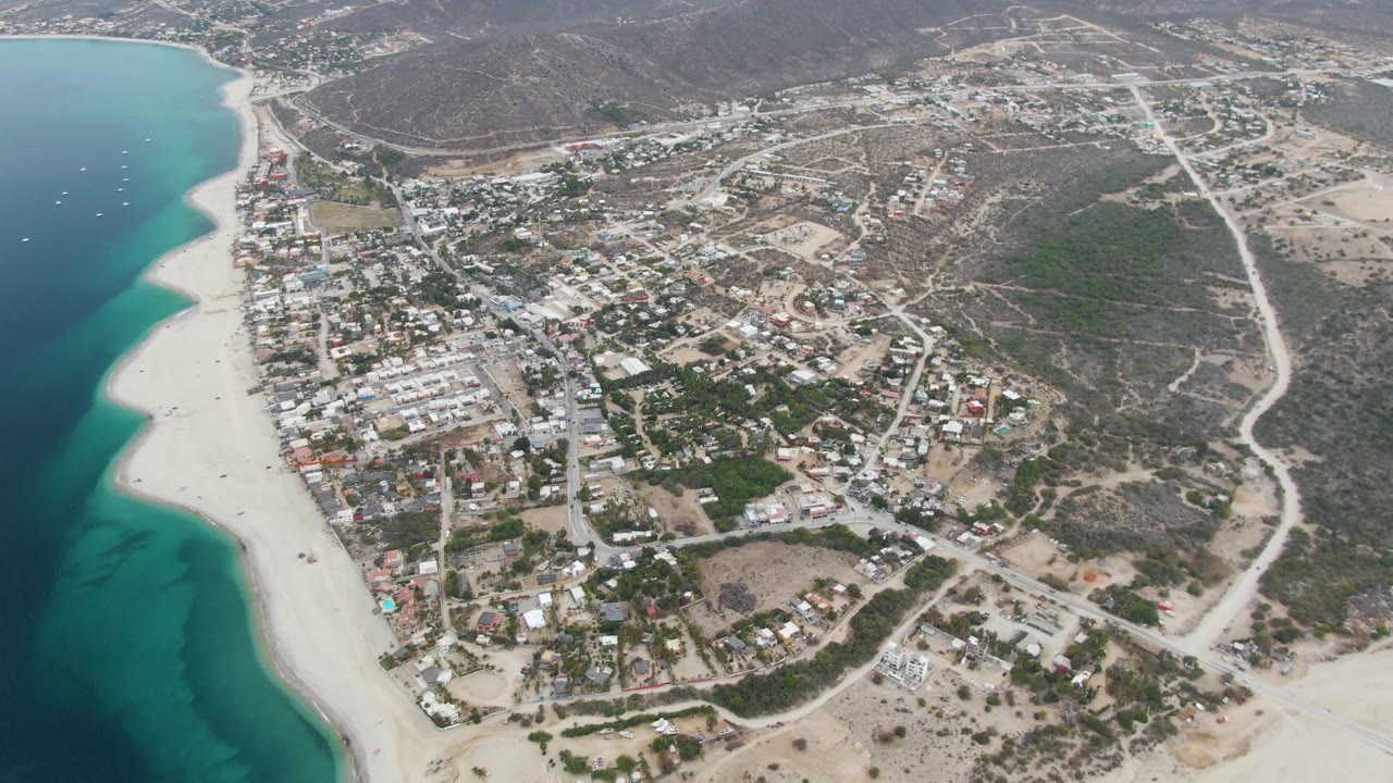 Opening aerial frame over Los Barriles, Baja California Sur, with turquoise ocean at left, pale beach, town buildings, dirt roads, dry hills, and no visible people.