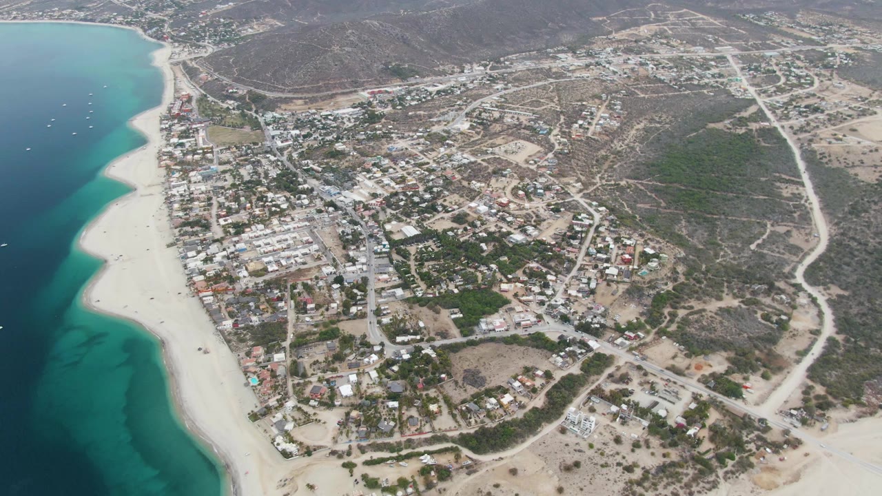 Mid-clip aerial frame of Los Barriles, with boats scattered offshore, a broad sand beach, low town buildings, pale roads across dry terrain, and no visible people.