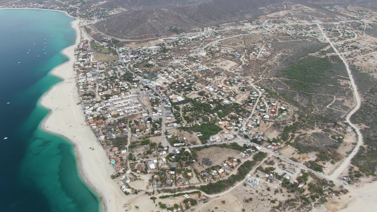 Aerial coastal town view with beach, water, boats, roads, and hills.