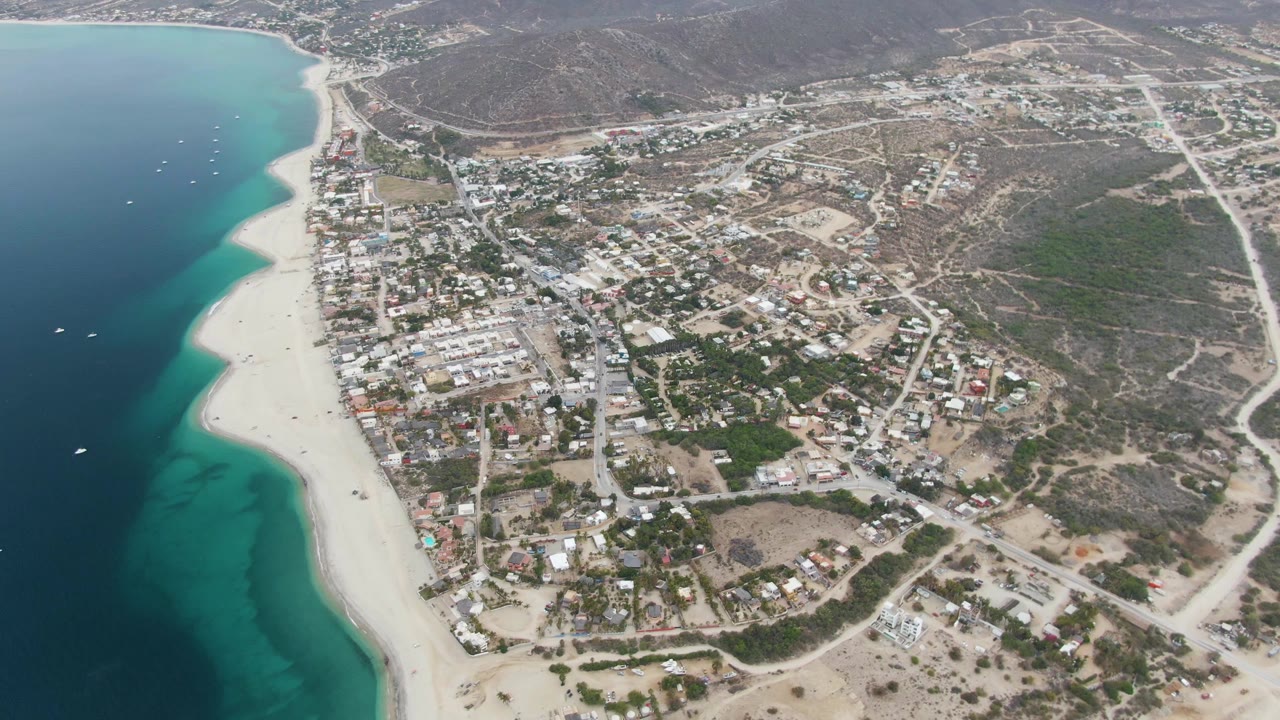 Opening aerial frame over Los Barriles, Mexico, showing a pale sandy beach along turquoise water, town roads and buildings inland, dry hills beyond, and no visible people.