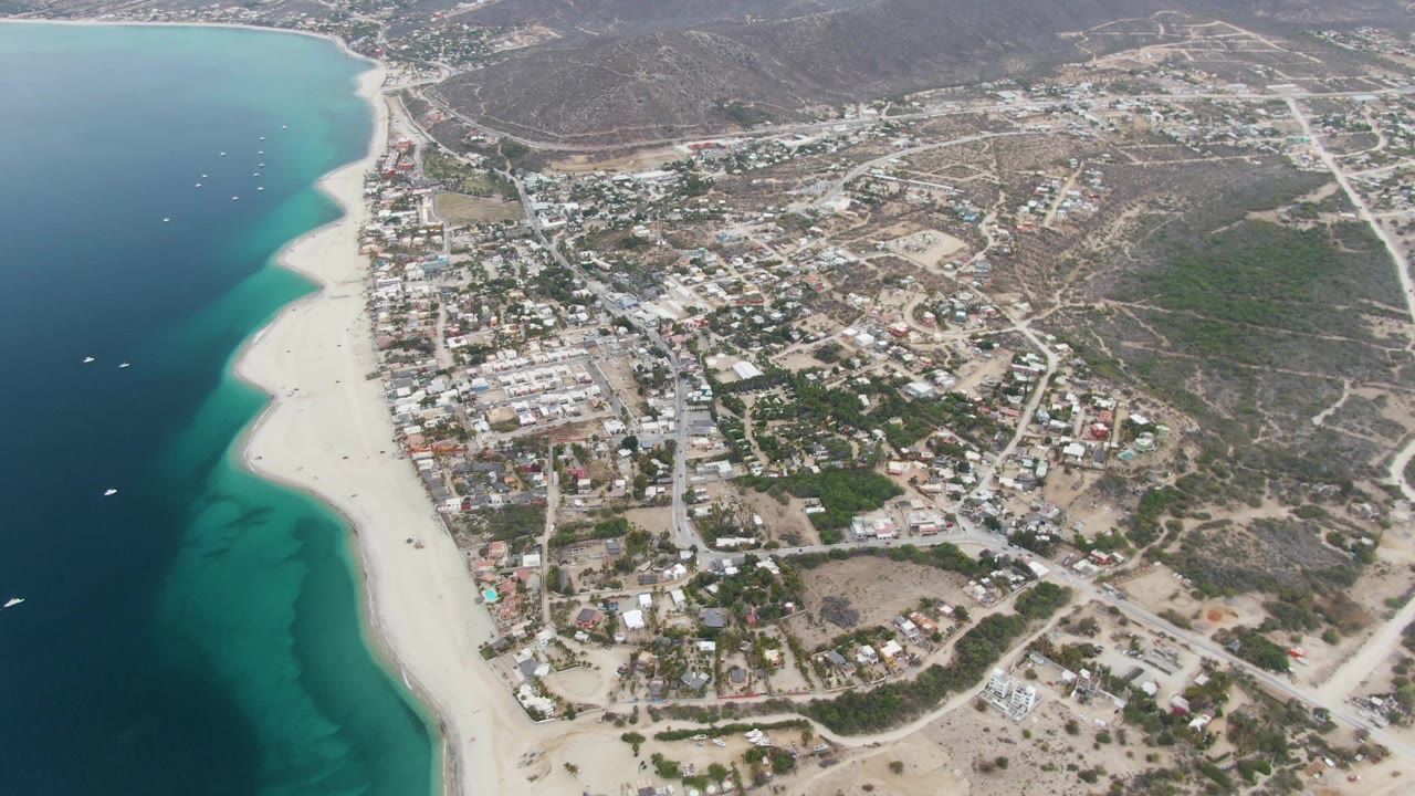 Second frame of Los Barriles coastline with the beach and turquoise shallows on the left, denser town blocks near center, inland roads, dry hillside terrain, and no visible people.