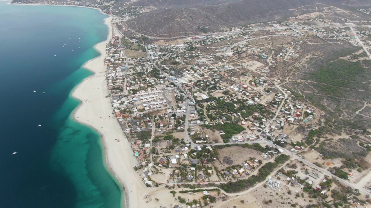 Aerial view along a long sandy beach with town buildings and hills inland.