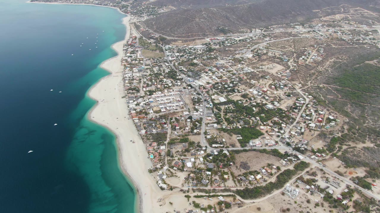 Aerial frame of Los Barriles showing deep blue ocean, turquoise shallows, a pale sandy beach, scattered small boats, town buildings, roads, dry hills, and tiny visible people on the shore.