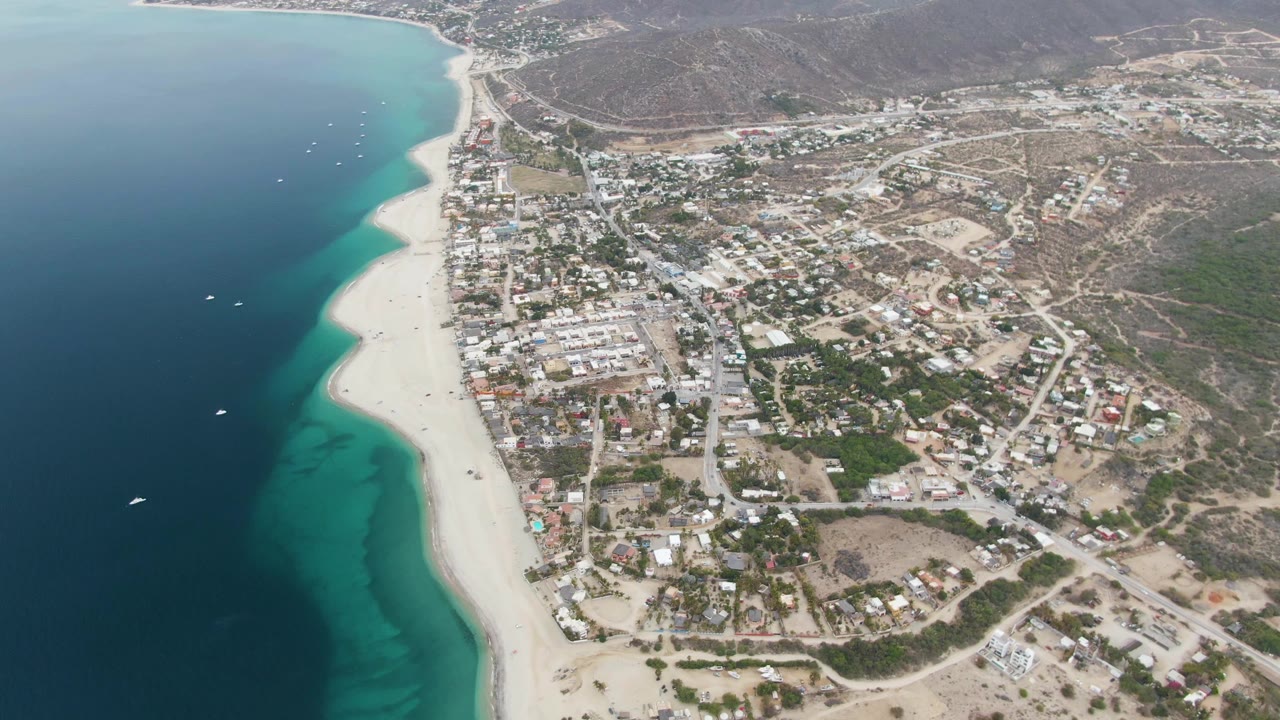 Los Barriles aerial frame with several small boats in the blue water, bright sand along the curved shore, a compact town inland, road intersections, scattered greenery, and distant dry terrain.