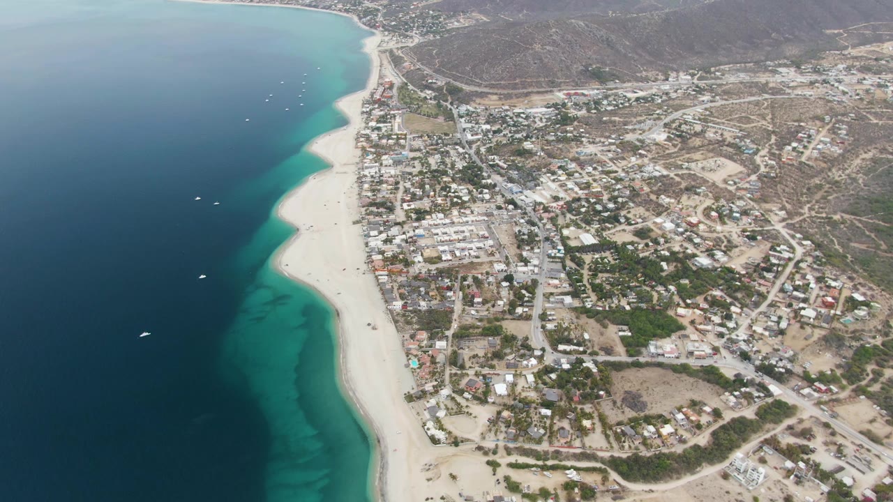 Aerial frame over Los Barriles, Mexico, showing deep blue ocean, turquoise shallows, a sandy shoreline, town buildings, dry hills, and no visible people at the start of the clip.