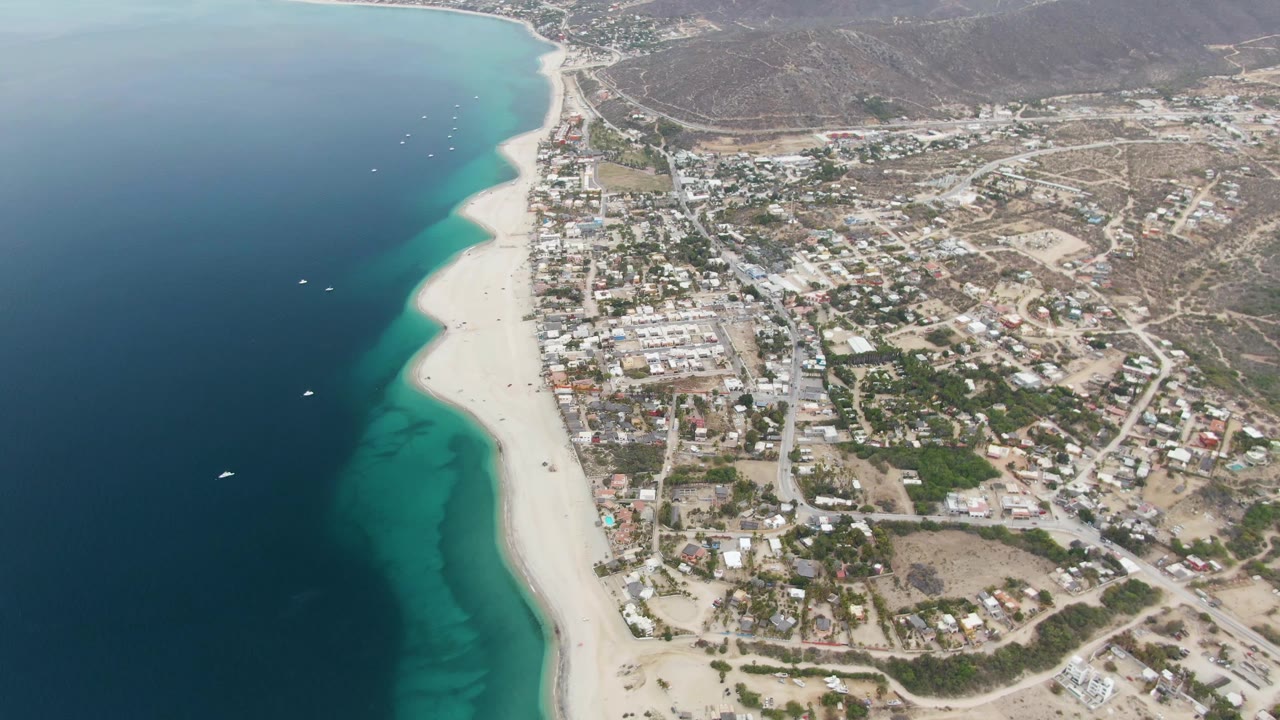 High aerial view of Los Barriles with turquoise water along the beach, scattered white boats offshore, dense low buildings inland, dry hills behind town, and no visible people.