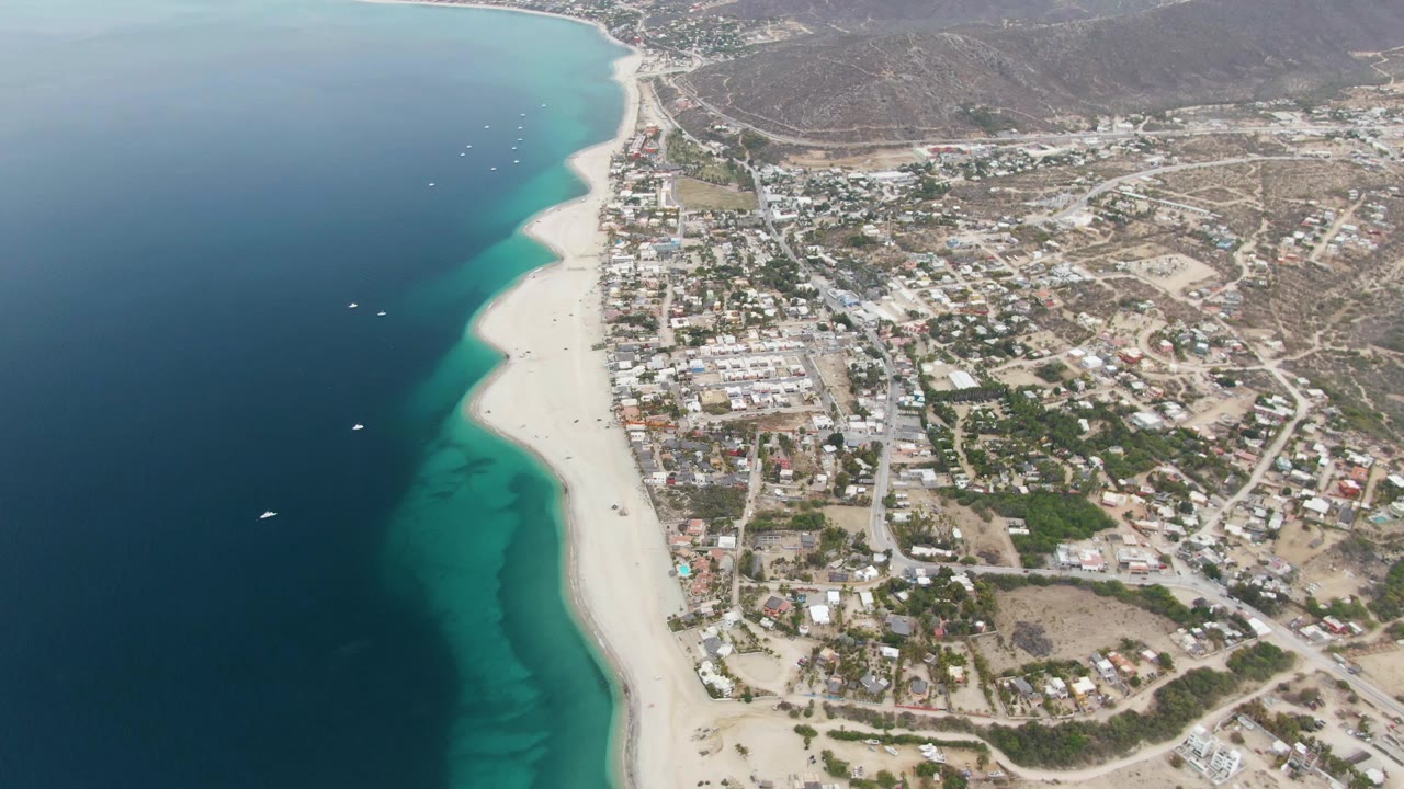 Drone aerial over Los Barriles, Mexico, with a long sandy beach dividing the blue ocean from low coastal buildings and arid hills, with no visible people.