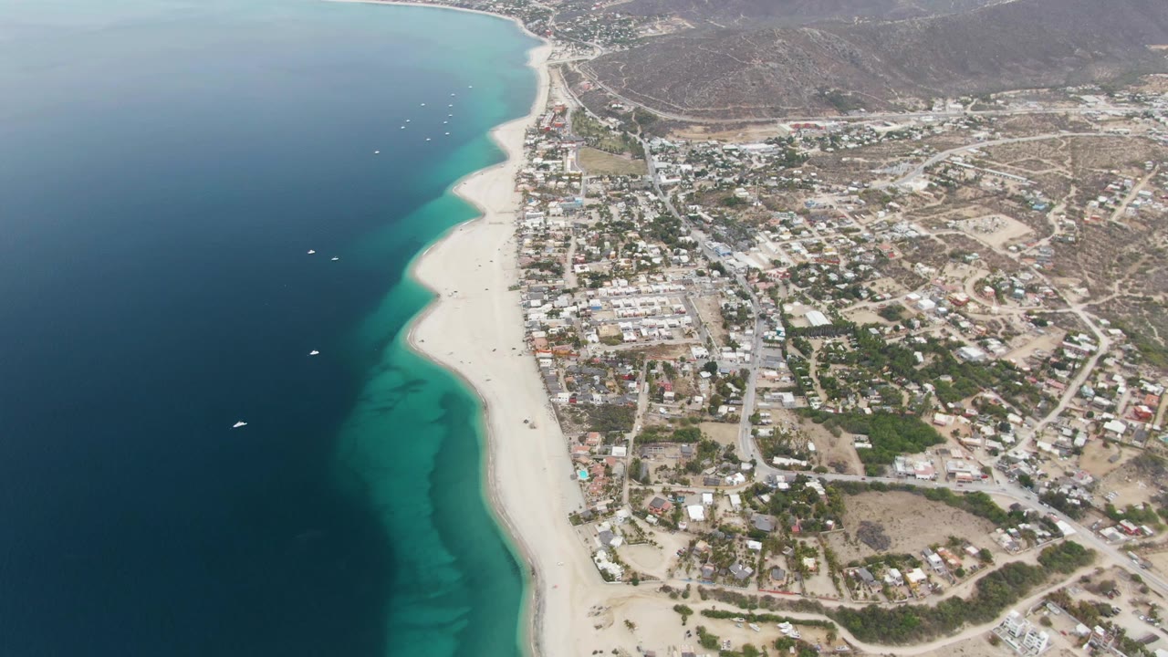 Aerial coastal town view with beach, boats, ocean, and hills.