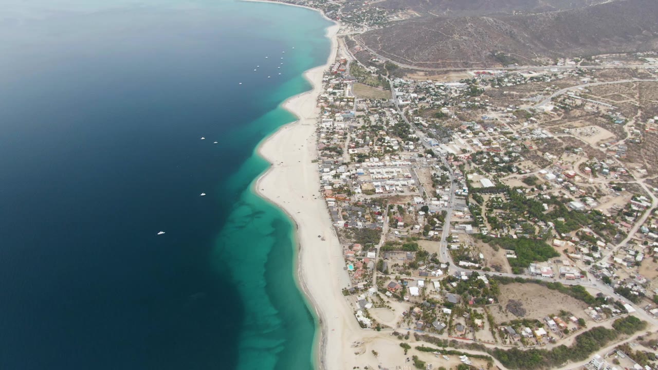 Aerial frame over Los Barriles, Mexico, showing deep blue ocean at left, turquoise shallows along a white sand beach, and town buildings and roads inland.