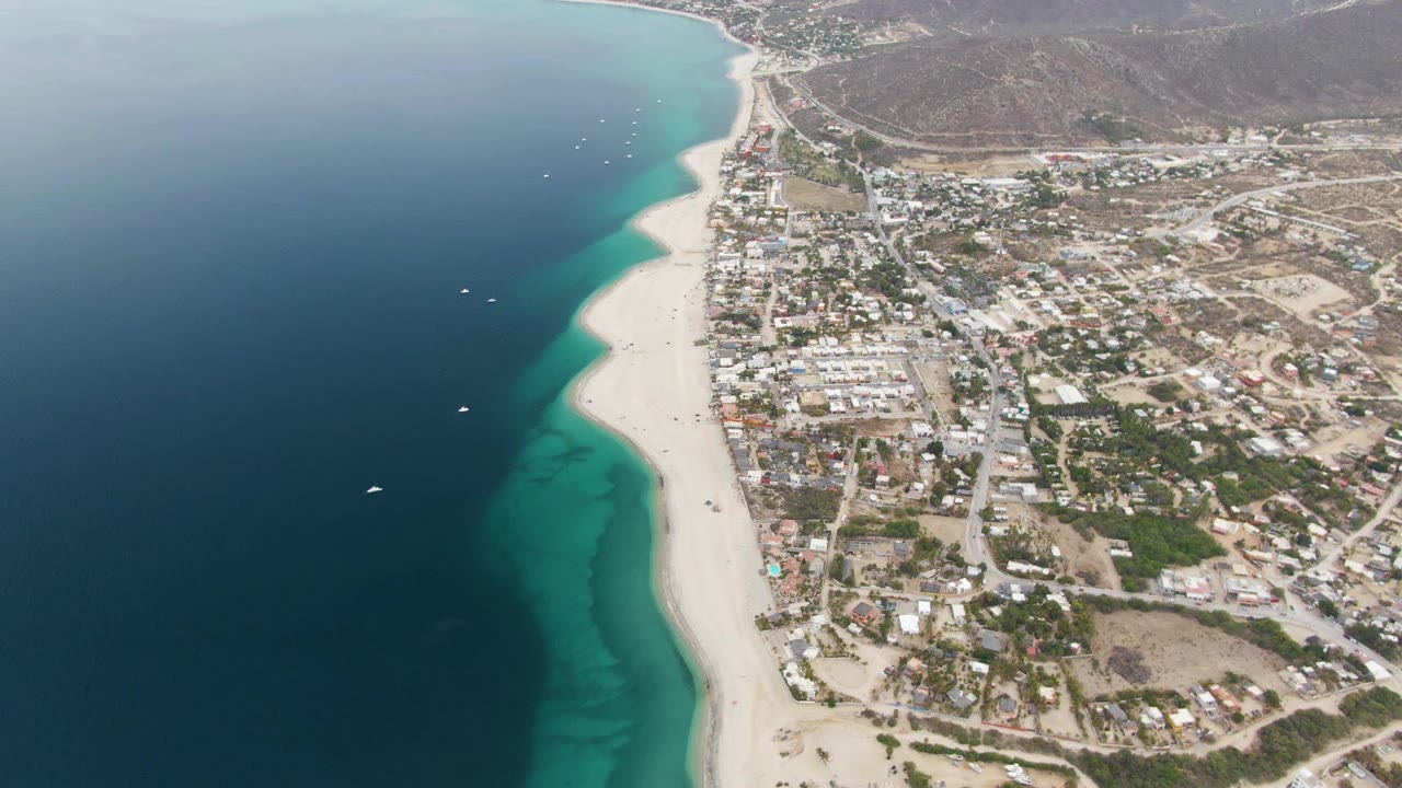 The frame shows Los Barriles from above with a stronger view of offshore boats in blue water, the pale beach edge, clustered buildings, roads, and dry hills behind the town.