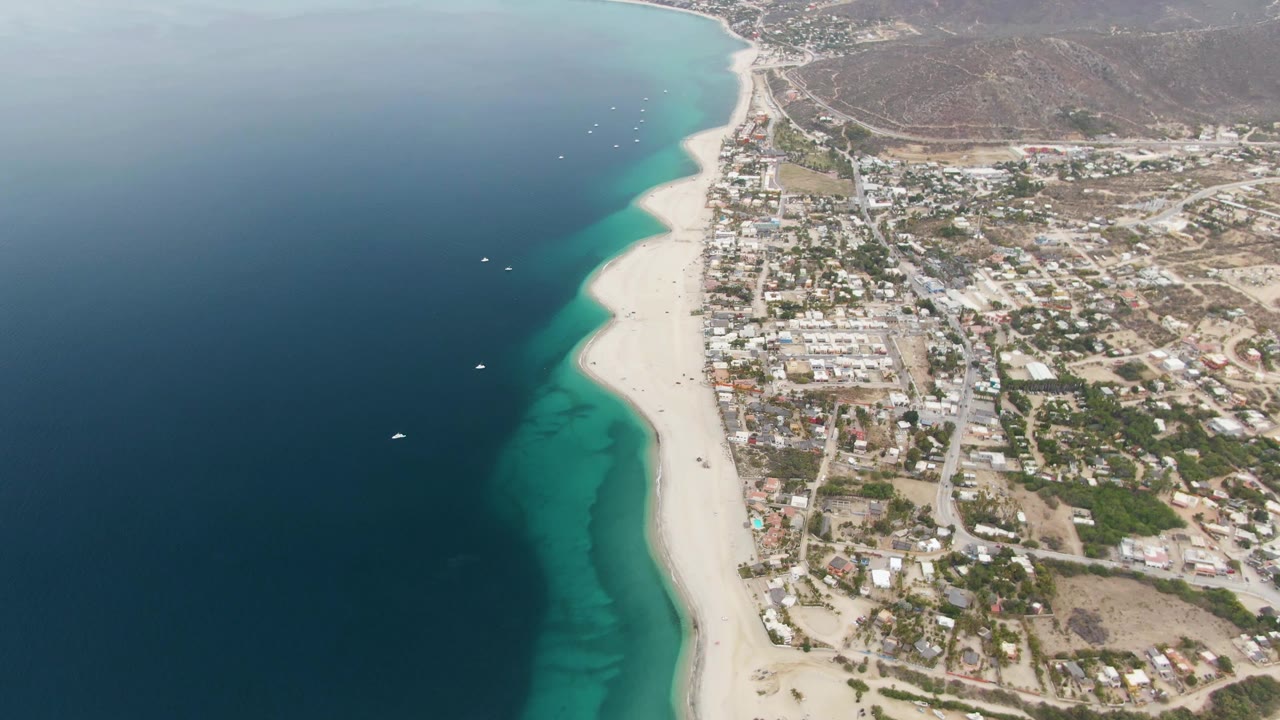 Aerial frame from Los Barriles with the beach curving up the coast, boats scattered in the water, compact town blocks beside the sand, and brown hills beyond.