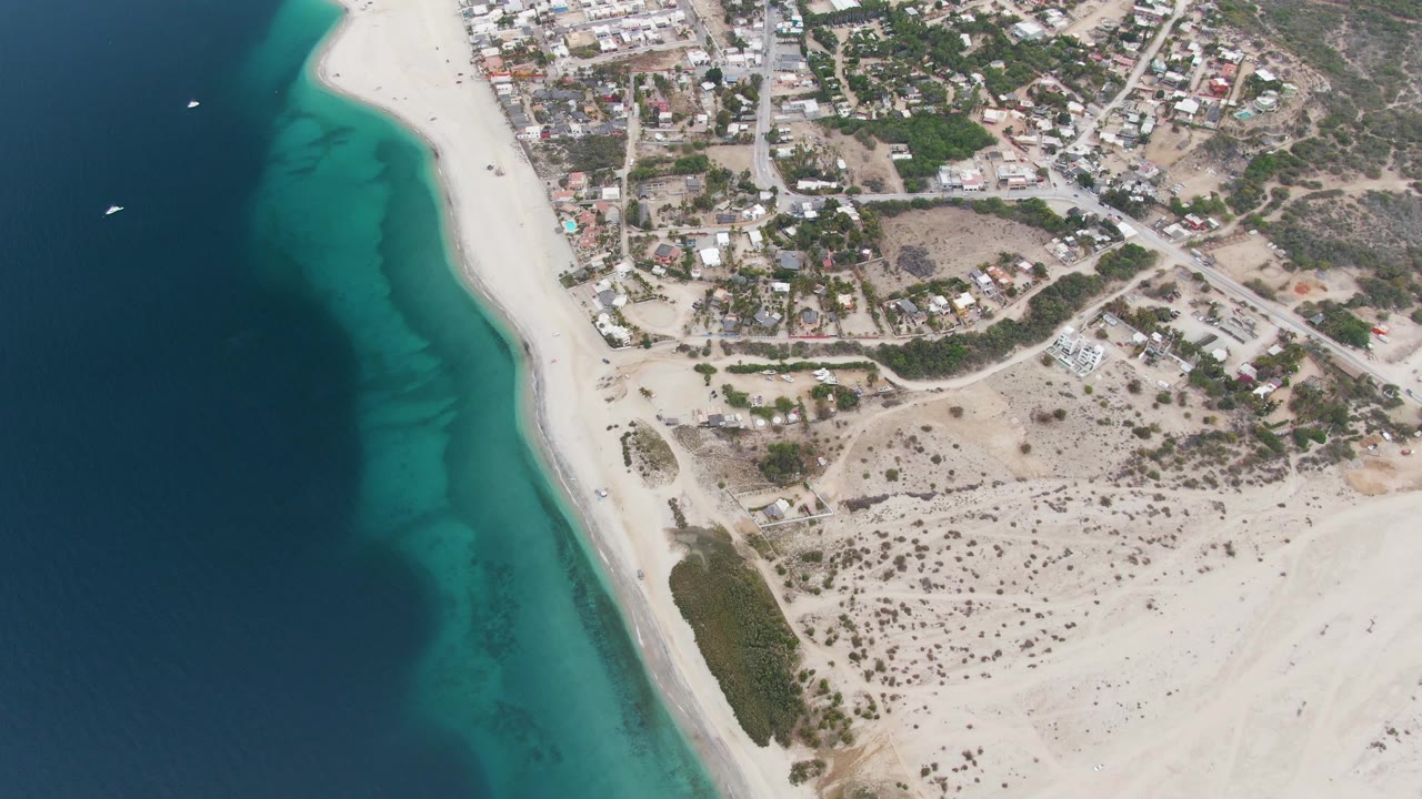 Overhead aerial frame of Los Barriles, Mexico with dark blue ocean, turquoise shallows, white sand, dry scrubland, town blocks, small offshore boats, and no visible people.