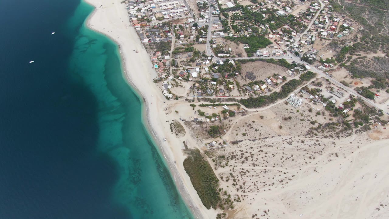 Drone view over Los Barriles showing the beach running diagonally beside turquoise water, with clustered town buildings inland, dry open lots, small boats offshore, and no visible people.