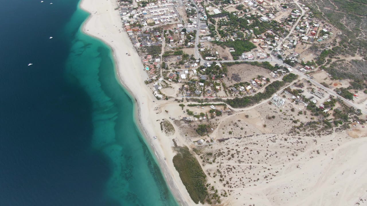 High aerial frame of Los Barriles with a broad strip of white beach, dark and turquoise ocean bands, small boats on the water, inland streets, homes, and no visible people.