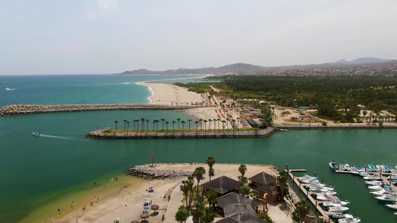 Aerial frame over Puerto Los Cabos Beach showing green marina water in front, a sandy beach with small visible people, palm-lined breakwaters, docked boats at right, and turquoise ocean beyond.