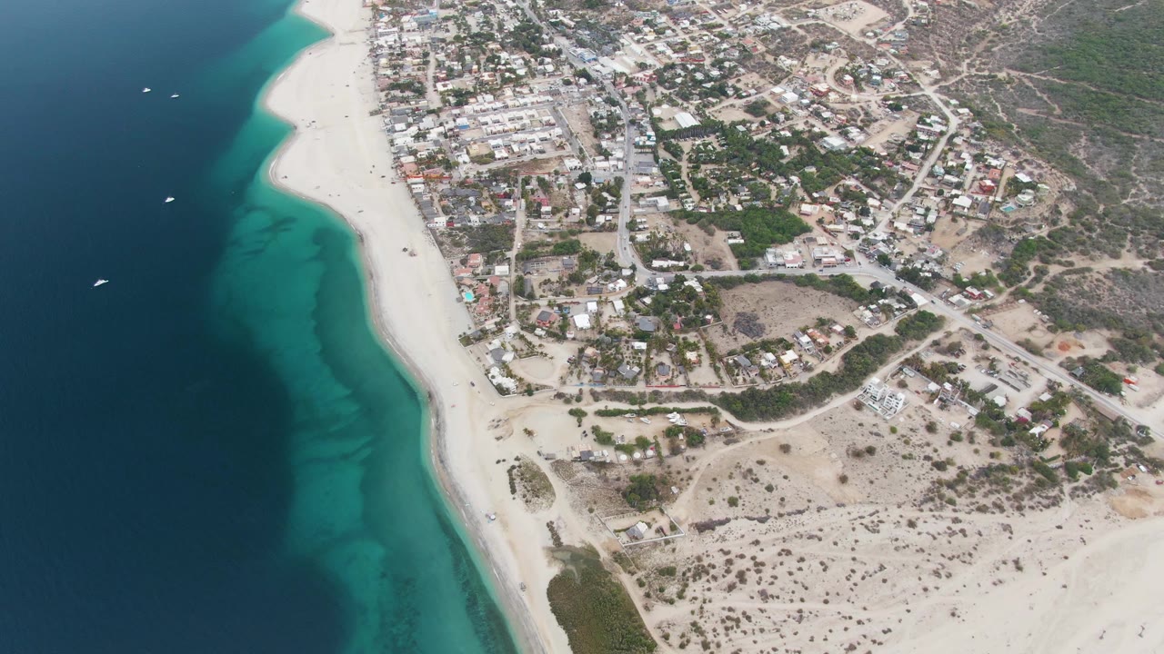 Opening aerial frame over Los Barriles, Mexico, with deep blue ocean, turquoise shallows, a white sand beach, coastal buildings, roads, dry terrain, and no visible people.