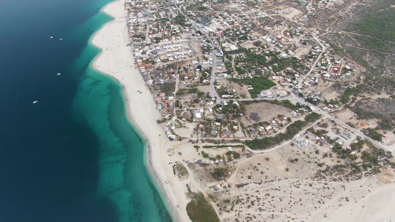Aerial frame over Los Barriles showing a brighter turquoise band along the beach, small boats offshore, clustered buildings, curving roads, sandy lots, and no visible people.