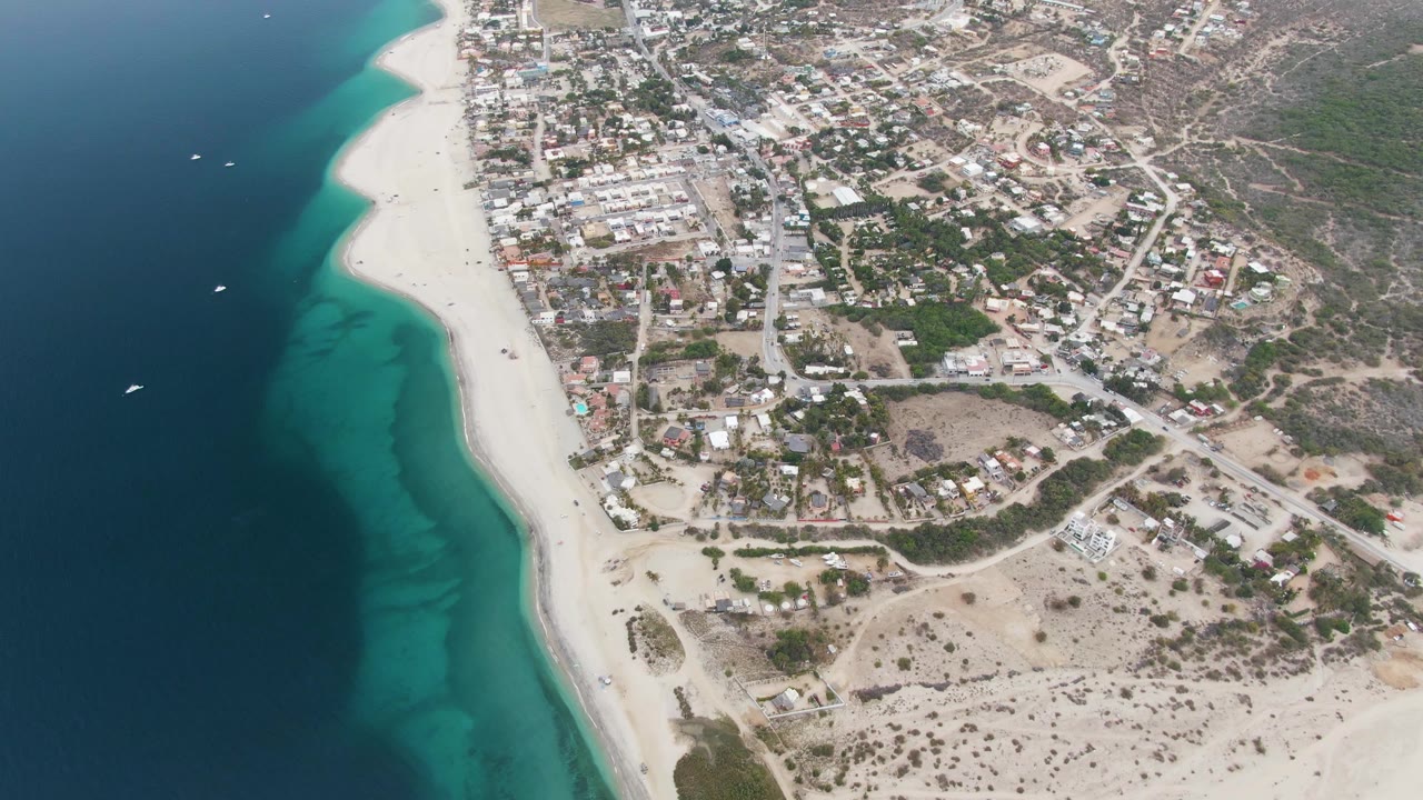 Aerial view of Los Barriles with several small boats on dark blue water, pale sand along the center, town buildings and road grids inland, scrubby hillsides, and no visible people.
