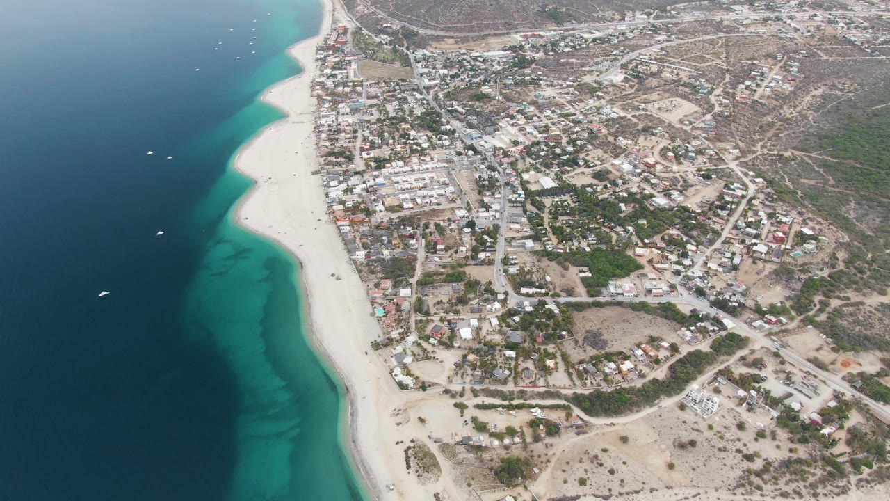 Aerial frame over Los Barriles showing deep blue ocean, turquoise shallows, a white sand beach, scattered boats, and low-rise town blocks inland.