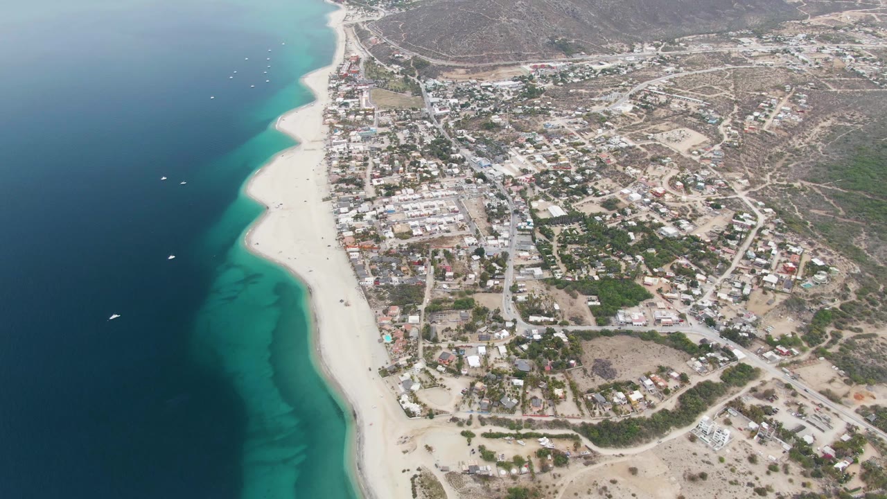 Los Barriles aerial frame with deep blue water on the left, turquoise bands near shore, visible beachgoers on the sand, boats offshore, and the town spreading toward arid hills.