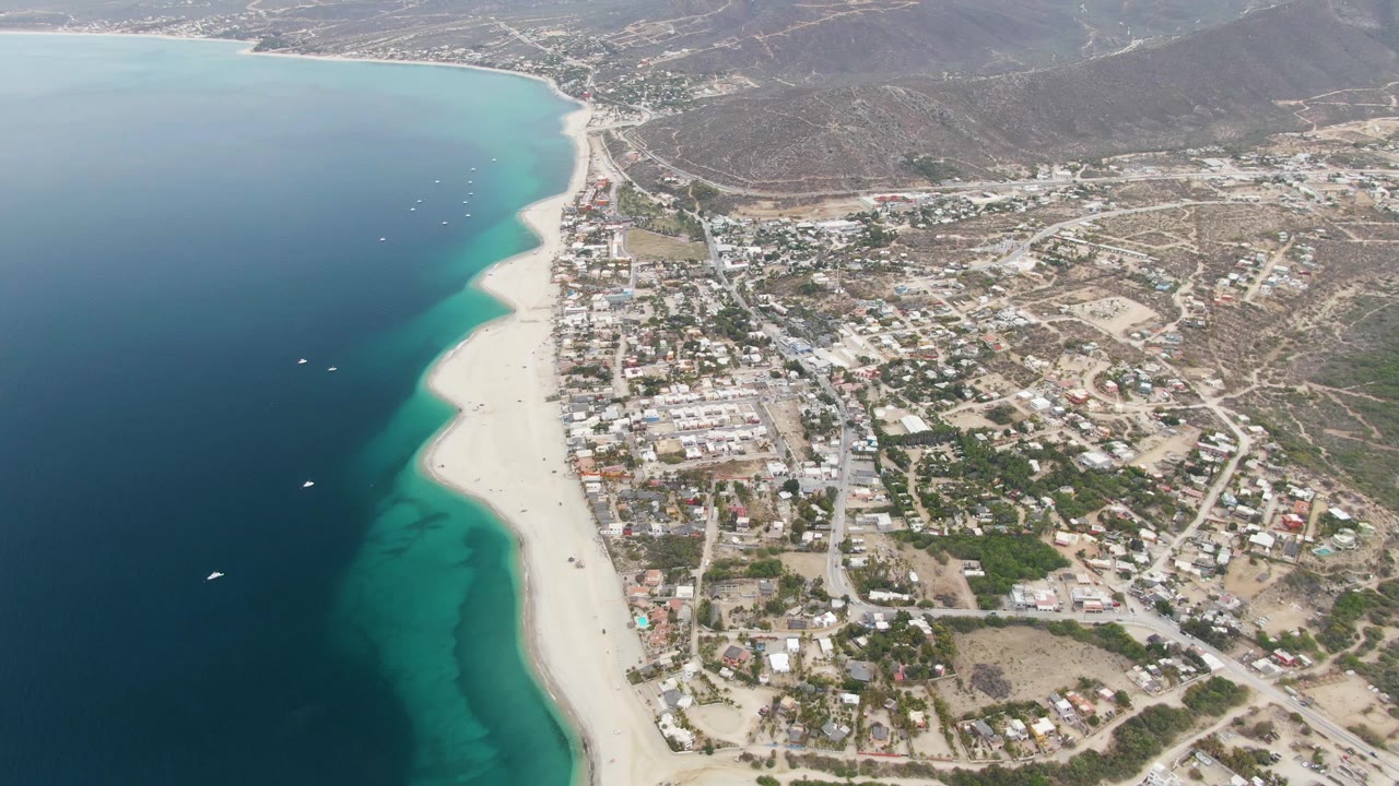 Opening aerial frame over Los Barriles, Mexico, with deep blue water at left, turquoise shallows along the sandy beach, town blocks inland, dry hills beyond, and no visible people.
