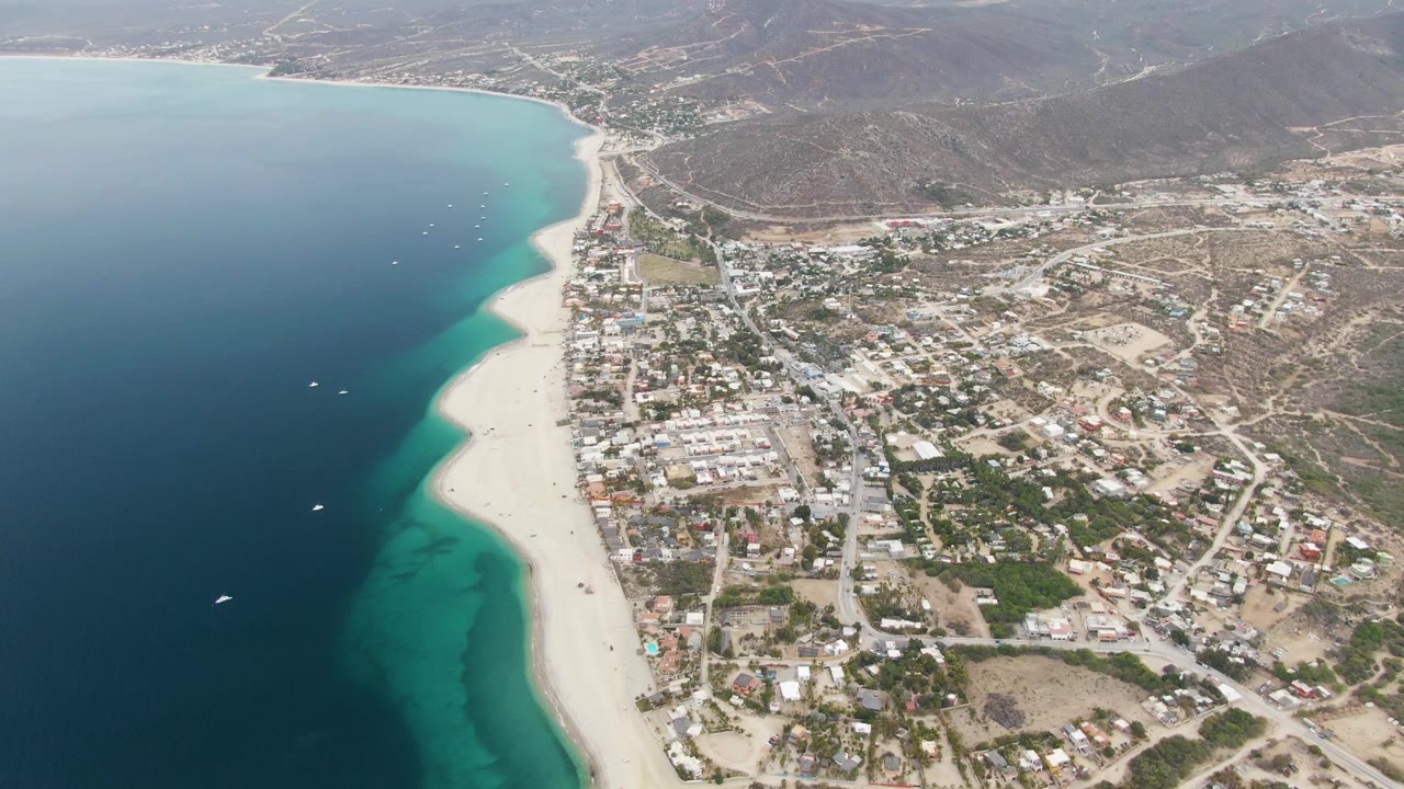Aerial frame over Los Barriles showing a long pale beach beside layered blue-green water, a cluster of low buildings and roads inland, mountains in the distance, and no visible people.