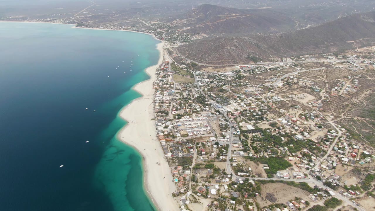 Mid-clip aerial frame in Los Barriles with the ocean occupying the left side, a bright sandy beach curving through the center, town streets and homes to the right, and no visible people.