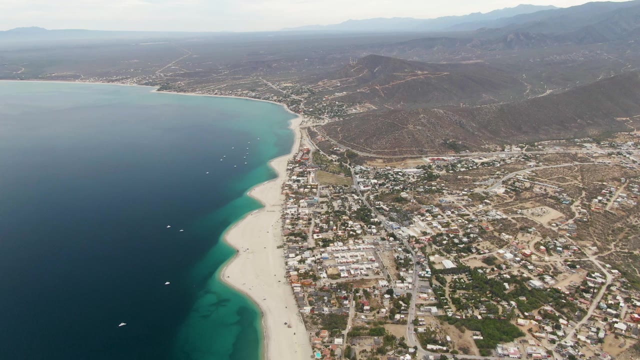 Aerial frame over Los Barriles, Baja California Sur, with deep blue water, turquoise shallows, sandy beach, town blocks, dry hills, distant mountains, and no visible people.