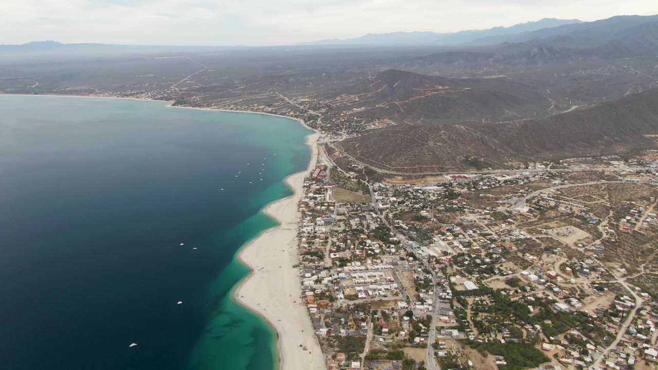 High aerial view of Los Barriles with the beach running down the center, dark blue ocean to the left, town streets to the right, mountains beyond, and no visible people.