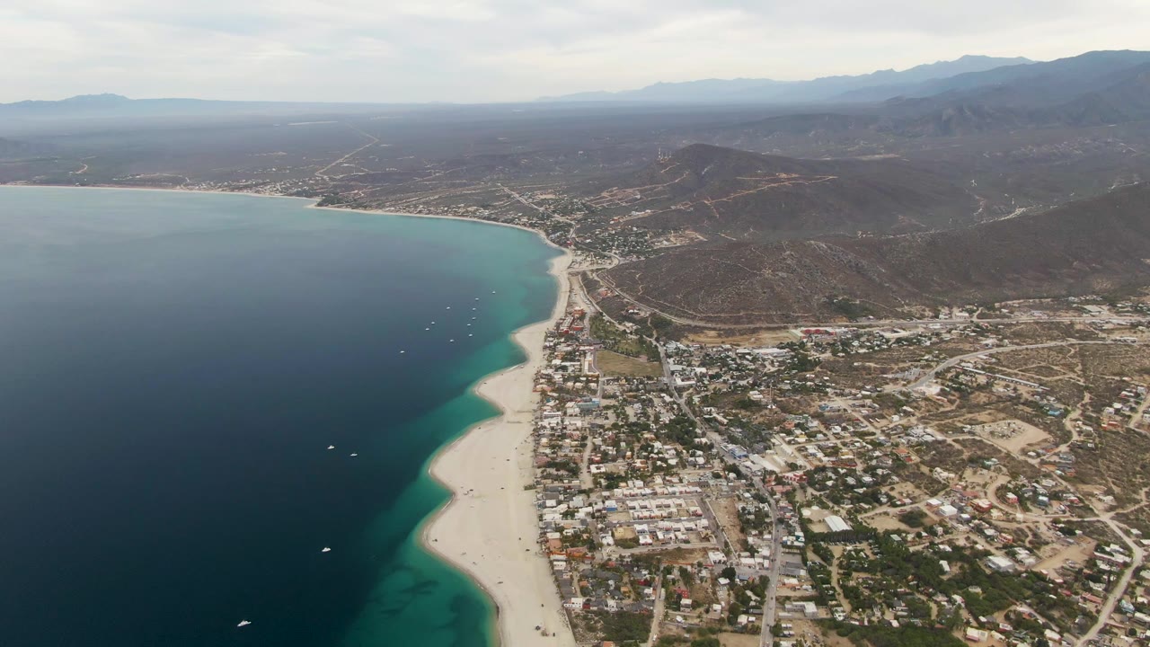 Wide Los Barriles aerial with more open water on the left, a curved pale beach through the middle, dense town blocks near the shore, dry inland terrain, and no visible people.