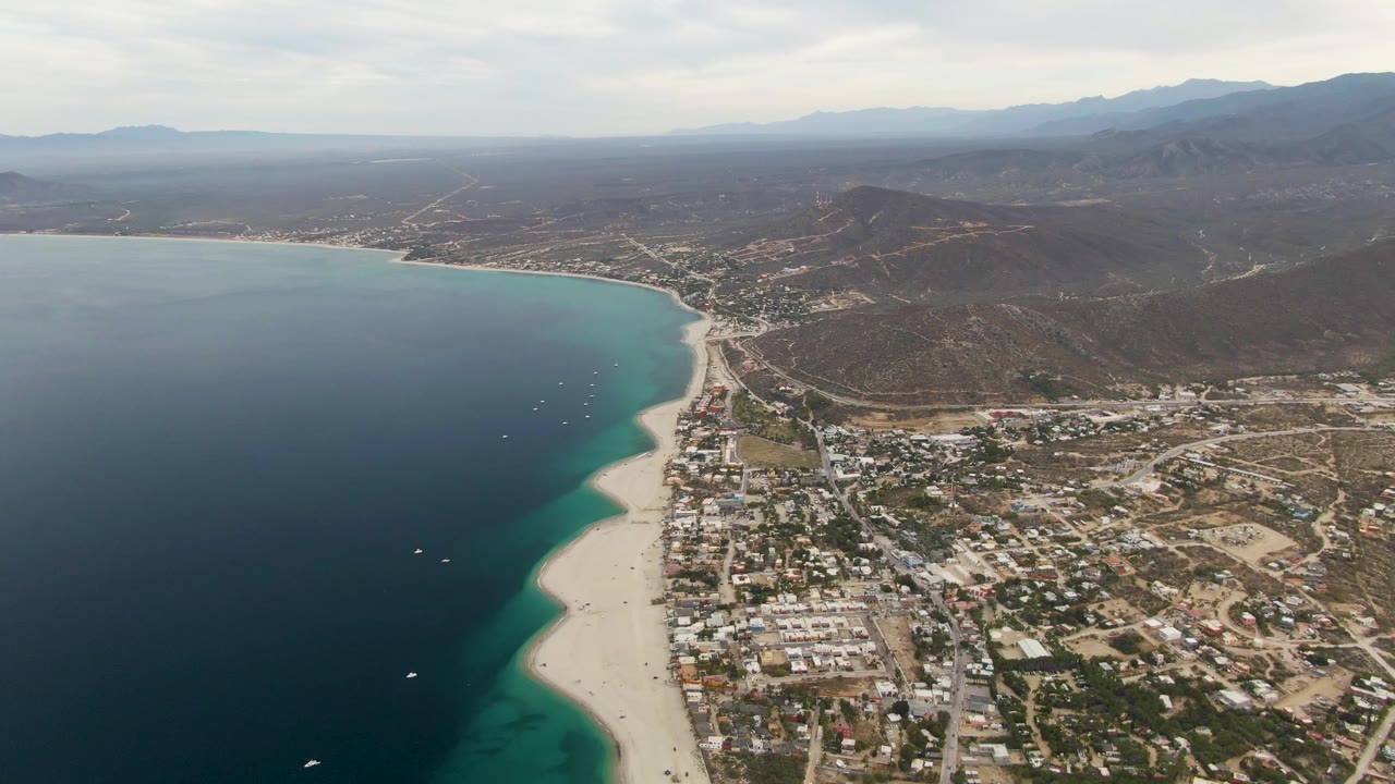 Aerial coast view with dark ocean, pale beach, town, and mountain backdrop.