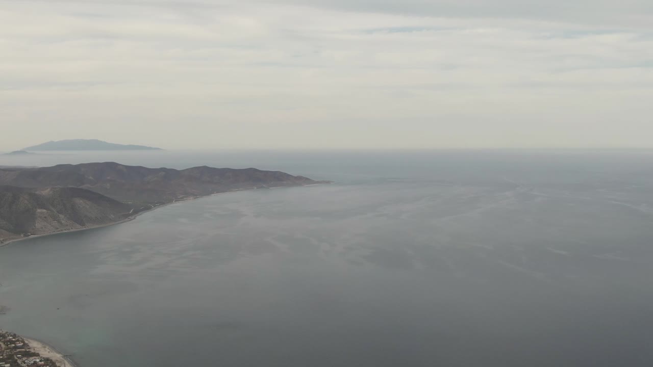 Aerial frame over Los Barriles showing gray-blue ocean, a sliver of shoreline at lower left, dark coastal mountains, a distant island shape on the horizon, and no visible people.
