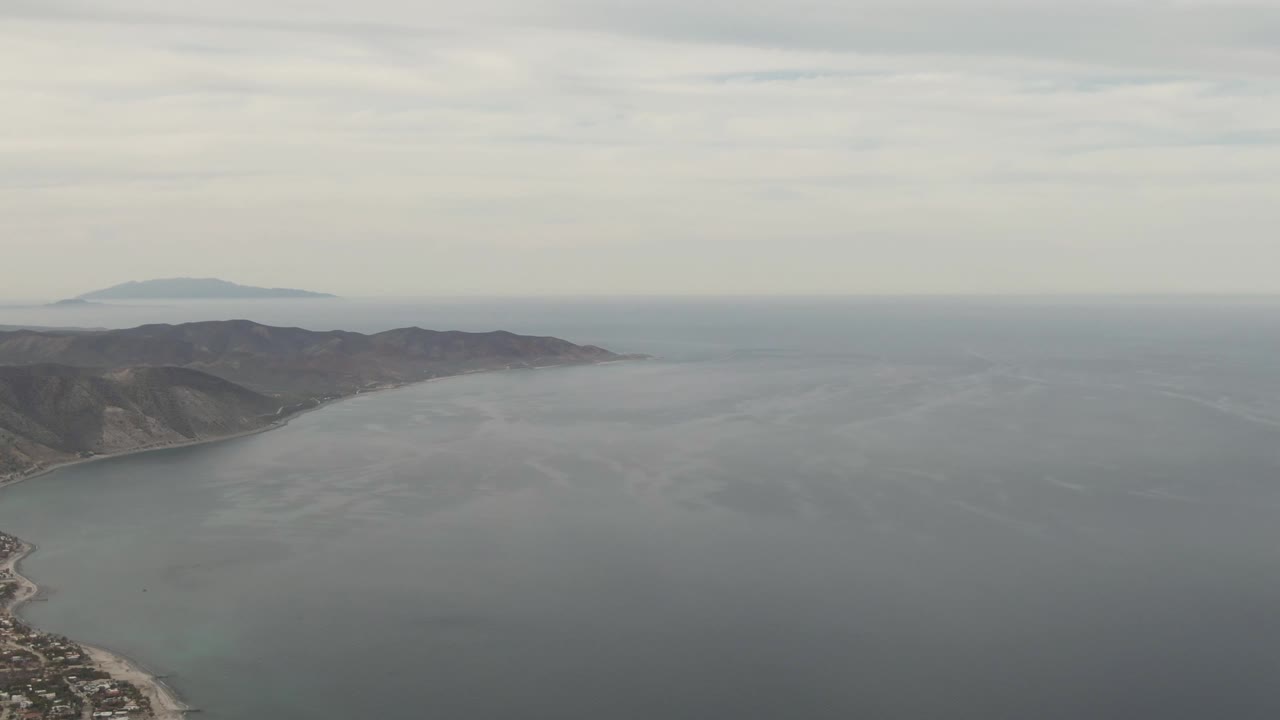 Aerial view of Los Barriles with more shoreline and town structures visible at the lower left, ocean across the foreground, mountains behind the coast, and no visible people.