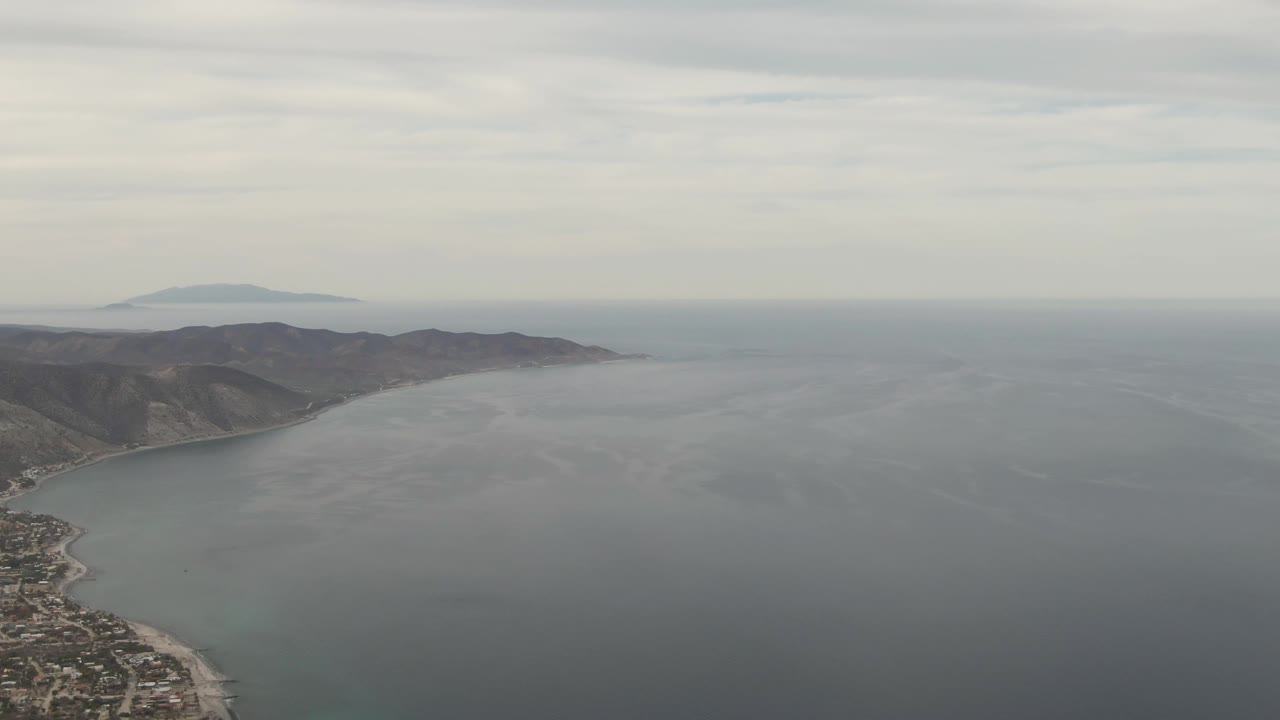 Wide Los Barriles aerial frame with the curved coast running down the left side, town blocks near the lower edge, broad ocean to the right, hazy mountains, and no visible people.